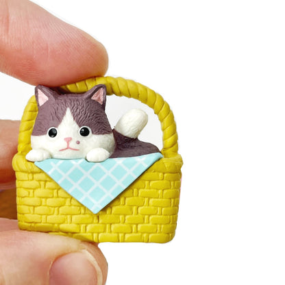 Fingers holding a cat figurine magnet of a kitten in a basket. 