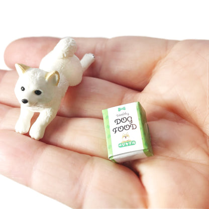 A hand holding a tiny white Shiba Inu toy dog figurine and a miniature cardboard dog food box.