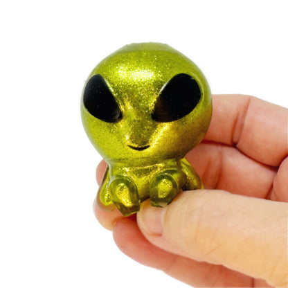 A hand holds a small squishy toy alien with glittery green skin and big black eyes.