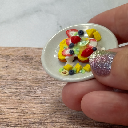 A hand holds a tiny pretend French toast on a mini ceramic plate.