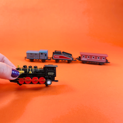 A hand holds a miniature pull back train engine while 3 other toy train cars are in the background on an orange backdrop 