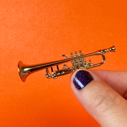 A hand with purple nail polish holds a miniature toy trumpet on an orange background