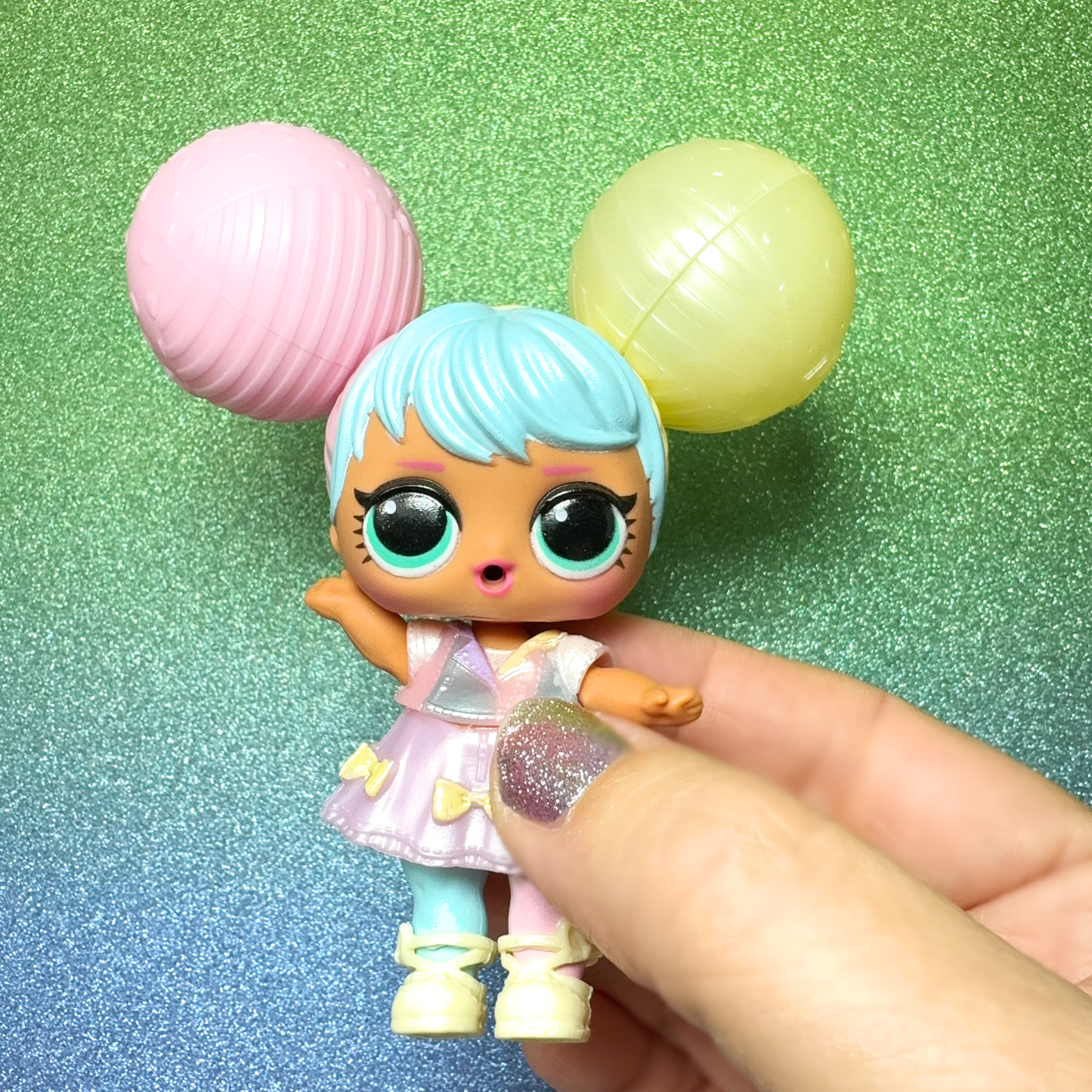 A hand holds an LOL Surprise Sooo Mini Doll against a sparkly green background