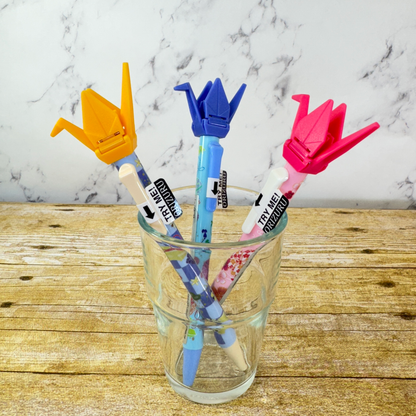 3 origami crane pens sit in a glass jar. They are pink, blue, and yellow.