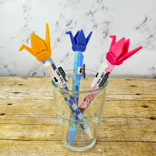 3 origami crane pens sit in a glass jar. They are pink, blue, and yellow.