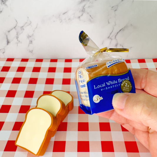 A hand holds 3 bread shaped erasers in a bread bag style packaging while 3 more bread shaped erasers lie on a red and white checkered tablecloth.