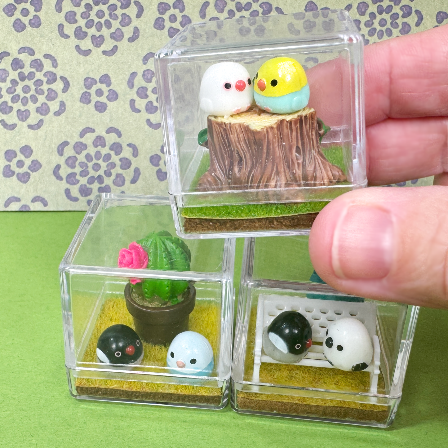 A hand holds a tiny bird terrarium figurine while two others are stacked underneath. 