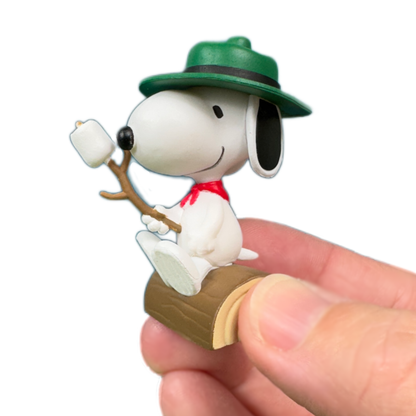 A hand holds a snoopy figure wearing a green hat and carrying a marshmallow on a roasting stick. The toy figure from Peanuts sits on a log. 