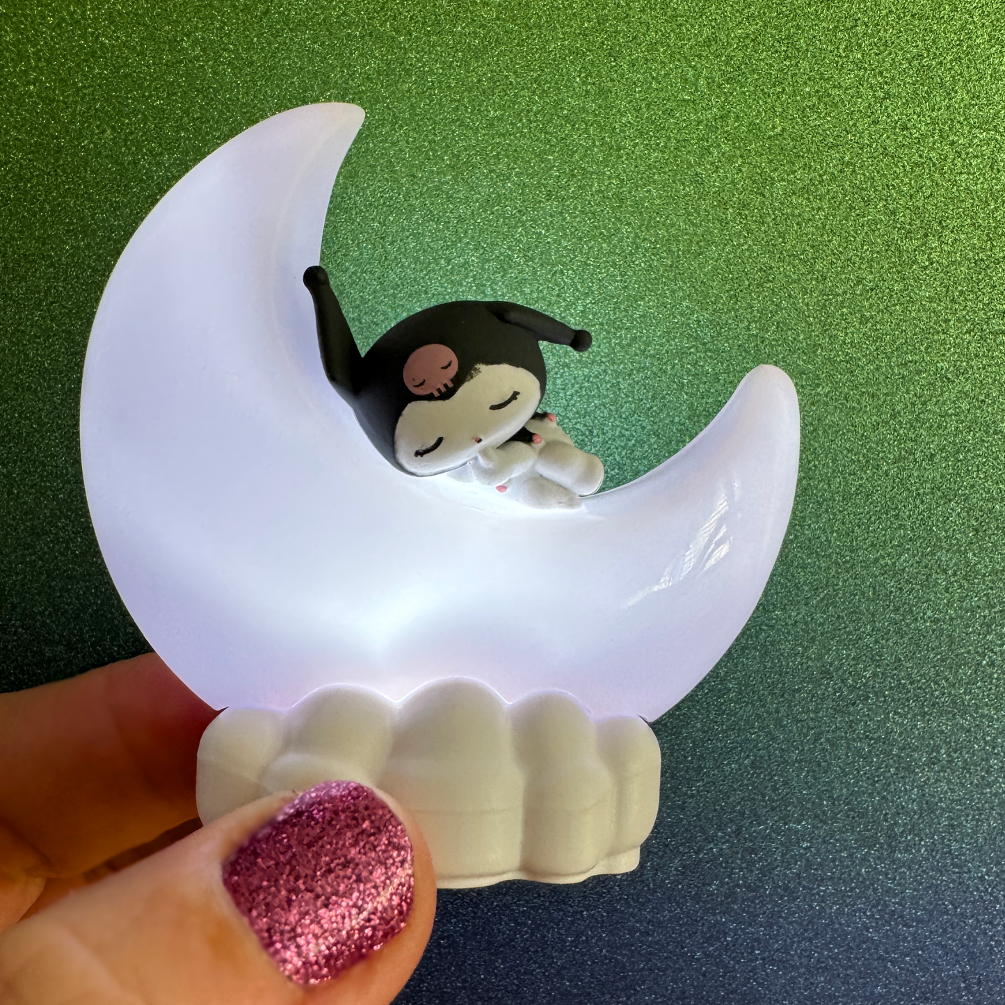 A capsule toy of a tiny figure of Sanrio's Kuromi character sleeping on a glowing moon shaped night light. 