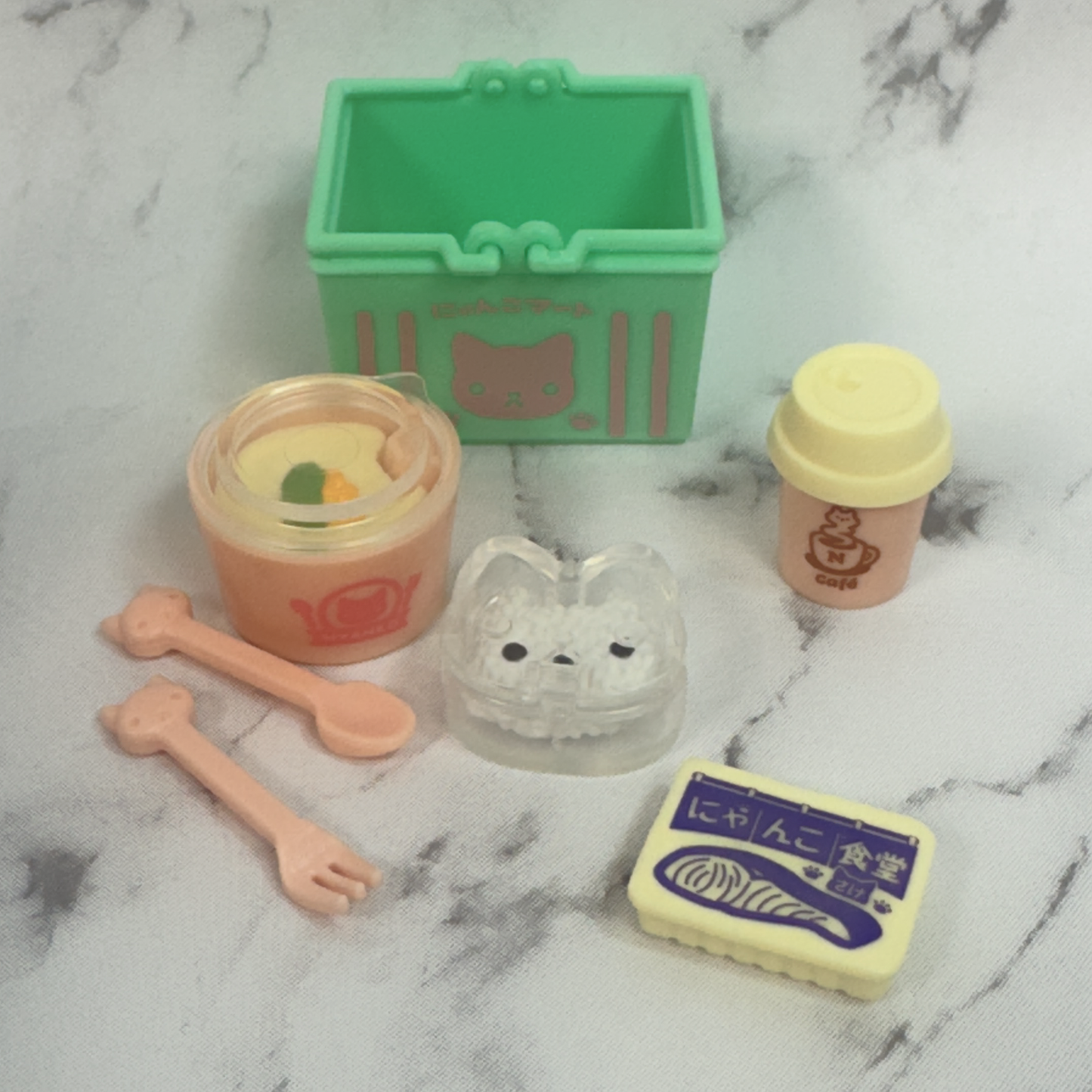 An opened capsule toy from the kitty cat market series. This toy includes a tiny aqua shopping basket, some pretend takeout foods, utensils, and a pink to-go coffee cup. Everything is cat themed.