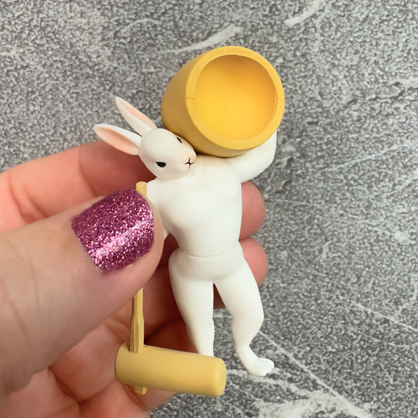 A hand holds a capsule toy rabbit figure with muscles modeling traditional mochi making techniques.