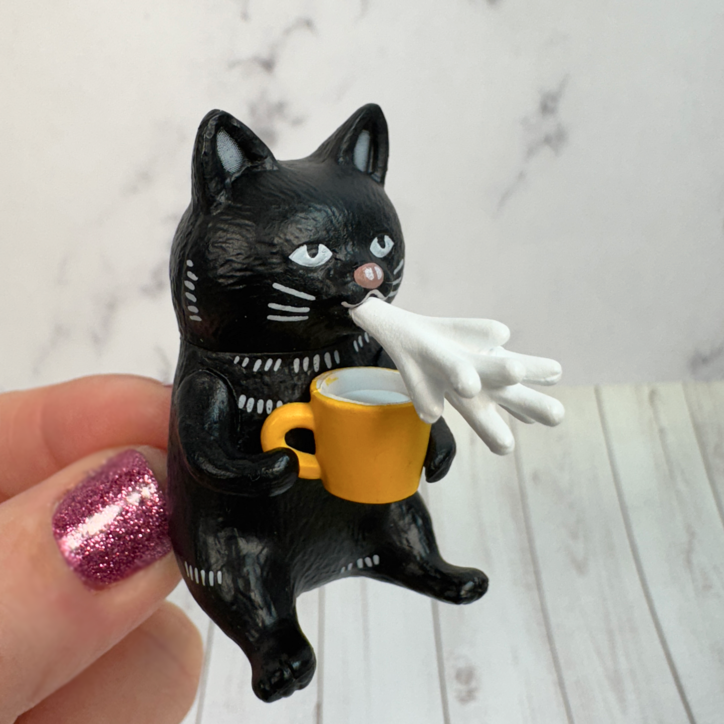 A hand holds a toy black cat figure spitting out milk while holding a yellow mug.