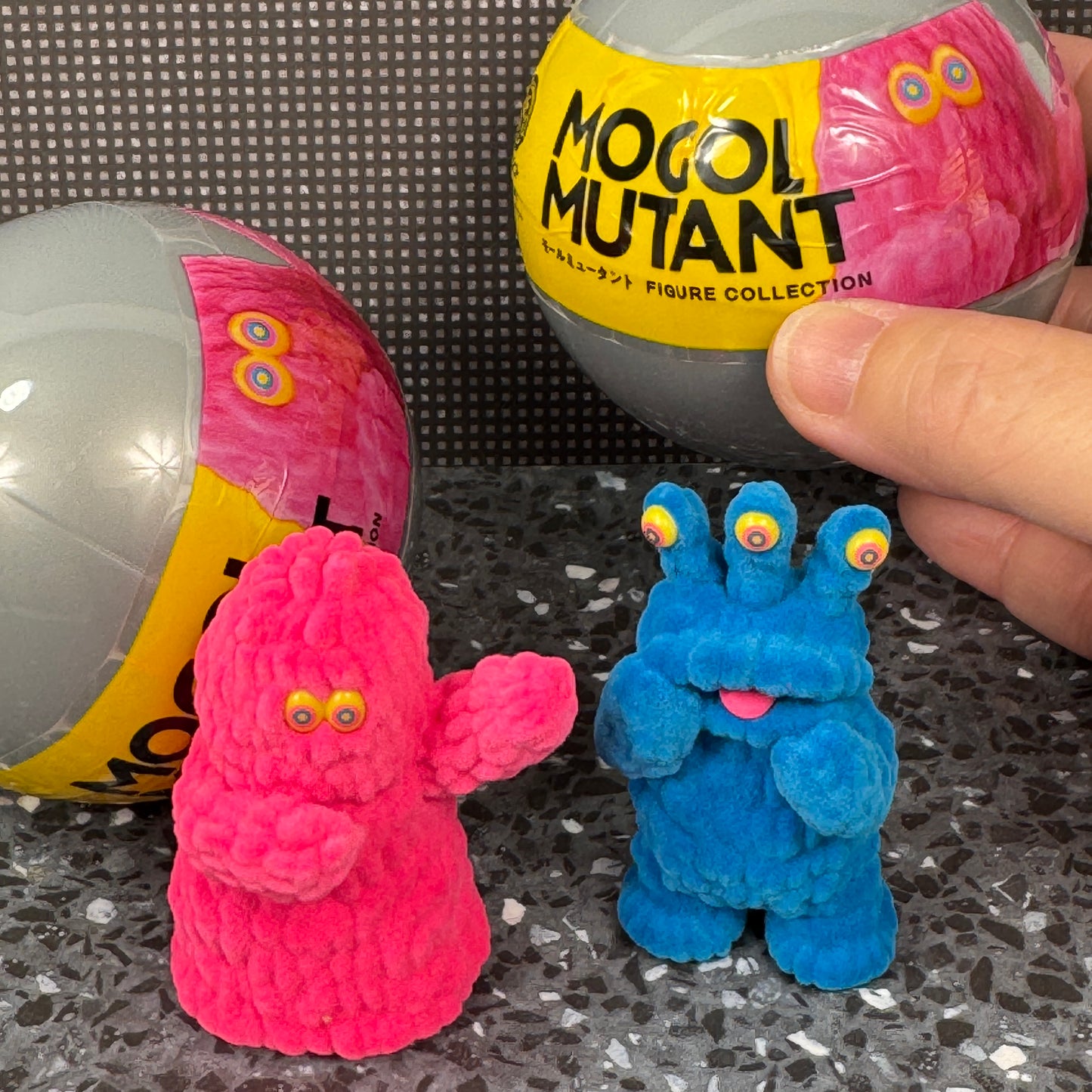 A hand holds a gachapon capsule from the Mogul Monster mall art figure collection while two fuzzy mutant monster figures stand in front.