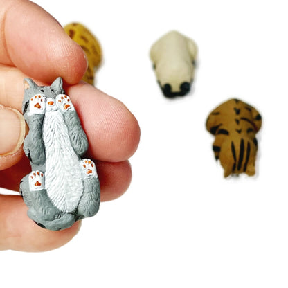 A hand holding a grey toy cat figurine with tiny paw prints. Other cat figurines are in the background.