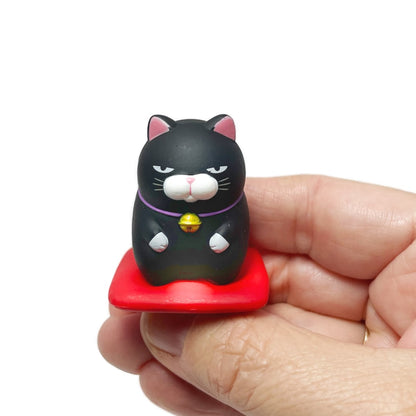 A hand holding a black toy cat figurine kneeling on a red cushion.