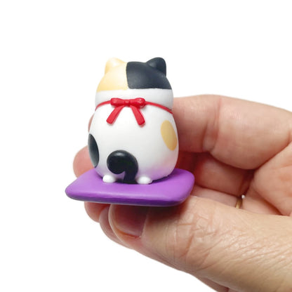 A hand holding a spotted toy cat figurine kneeling on a purple cushion. The back of the cat is shown with a red bow, fluffy tail, and cute feet.