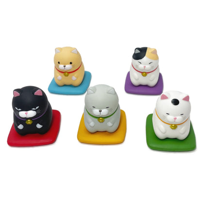 A group photo of five toy cat figurines kneeling on cushions, each with a unique color and design.